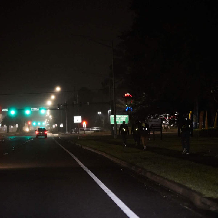 A nighttime scene shows a dimly lit road with a car driving toward a traffic light. Three people, wearing dark clothing and GORUCK Reflective Ruck Bands for nighttime visibility, walk in single file on the sidewalk. The street is illuminated by overhead lights, and the sky is overcast.