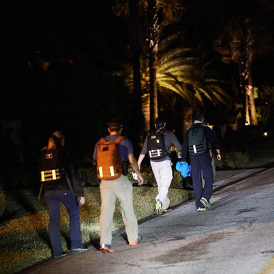 Four individuals walk along a dimly lit street at night, their backpacks outfitted with GORUCK Reflective Ruck Bands to enhance their visibility. They appear to be making their way toward a tree-lined, low-light area, possibly in a park or residential neighborhood.