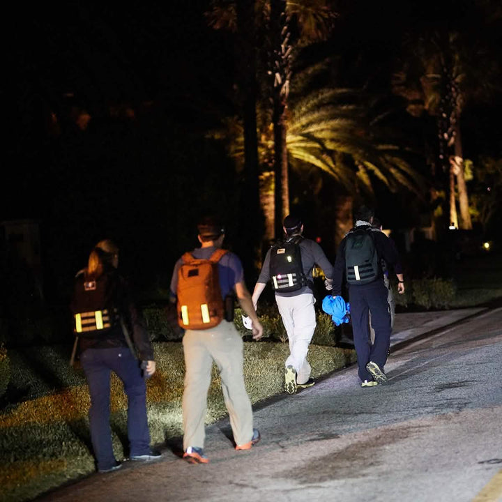 Four individuals walk along a dimly lit street at night, their backpacks outfitted with GORUCK Reflective Ruck Bands to enhance their visibility. They appear to be making their way toward a tree-lined, low-light area, possibly in a park or residential neighborhood.