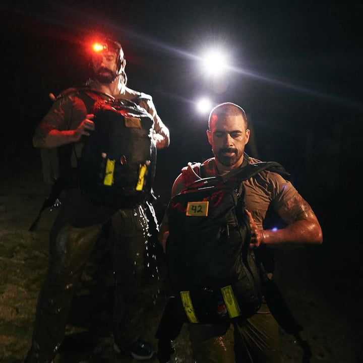 Two individuals wearing headlamps navigate the night with large, numbered backpacks. Despite being wet and weary, their gear is enhanced by GORUCK's Reflective Ruck Bands with MOLLE webbing for added versatility. A bright light in the background boosts nighttime visibility, casting a dramatic glow over the scene.