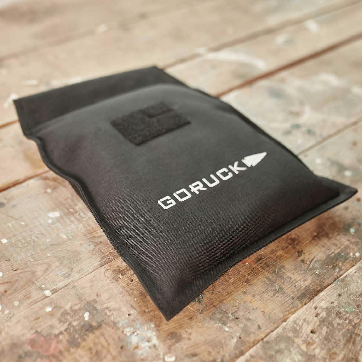 A black Ruck Sandbag - 10LB from GORUCK with a VELCRO® closure rests on a rustic, weathered wooden surface. This rectangular sandbag showcases the GORUCK logo in white and includes a velcro patch at the top, enhancing both durability and style in its rugged design.
