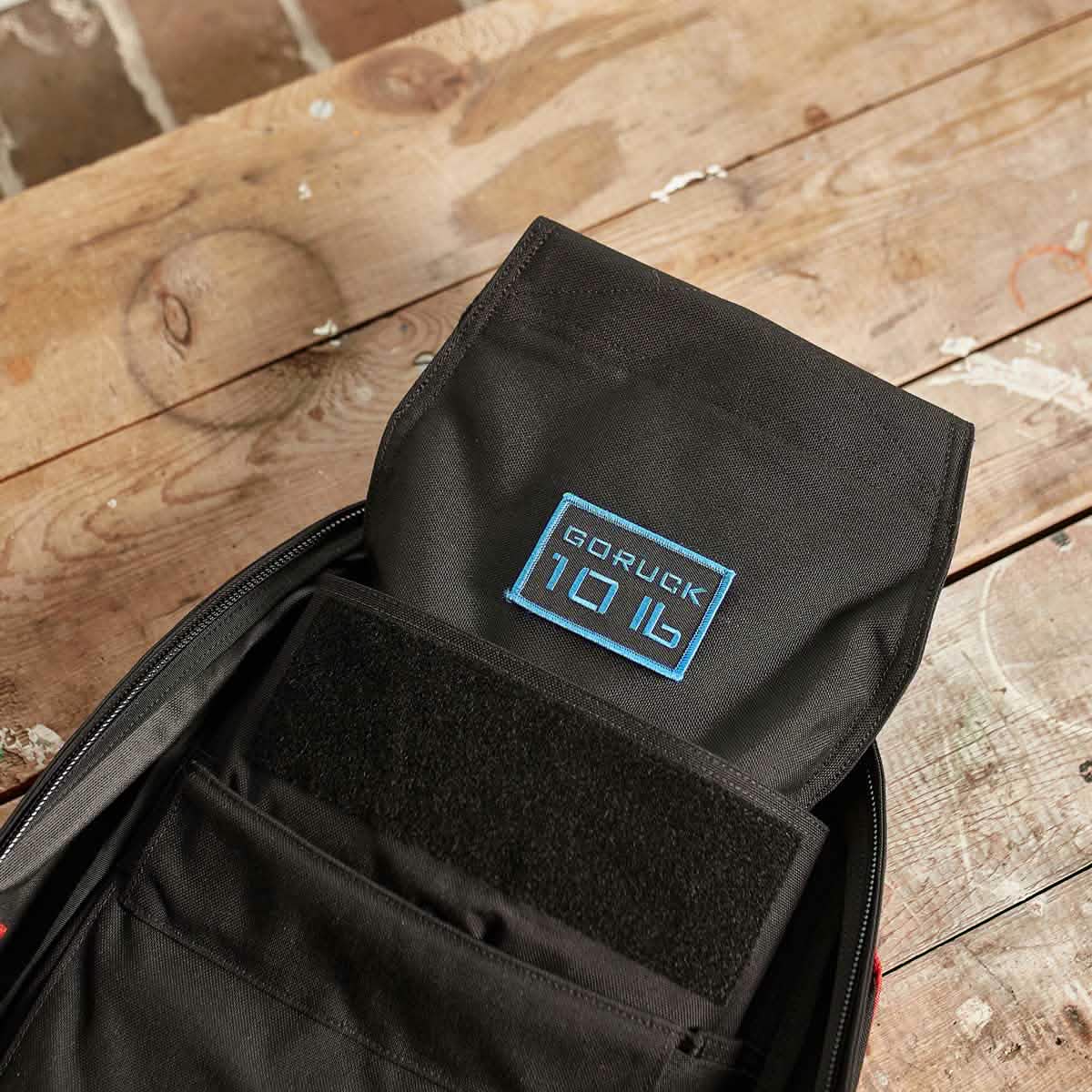 A black Ruck Sandbag - 10LB by GORUCK, featuring a VELCRO® closure, sits open on a rustic wooden table, revealing its interior. Inside, you can see the distinctive blue GORUCK logo against the backdrop of its sleek black material. The table holds character with white paint marks and scratches.