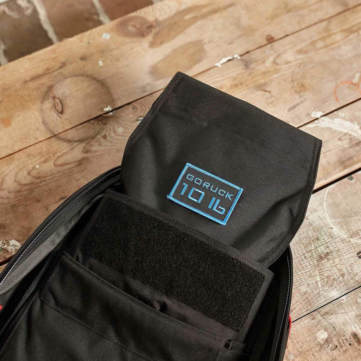 A black Ruck Sandbag - 10LB by GORUCK, featuring a VELCRO® closure, sits open on a rustic wooden table, revealing its interior. Inside, you can see the distinctive blue GORUCK logo against the backdrop of its sleek black material. The table holds character with white paint marks and scratches.