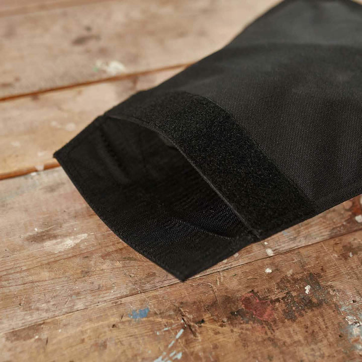 Close-up of a black fabric sleeve with VELCRO® closure on a wooden table. The sturdy fabric, characteristic of the GORUCK Ruck Sandbag - 10LB, suggests suitability for protective or industrial use. The table surface shows signs of wear and paint splatters.