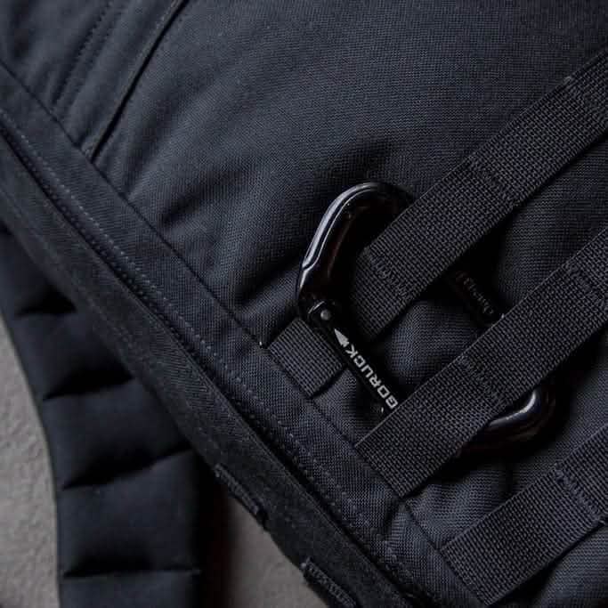 Close-up of a black backpack highlighting robust black straps and a small center clip. The textured fabric incorporates climbing-certified hardware, including SMC Gear's Carabiner with the GORUCK logo, while a brand label is partially visible on one of the straps.