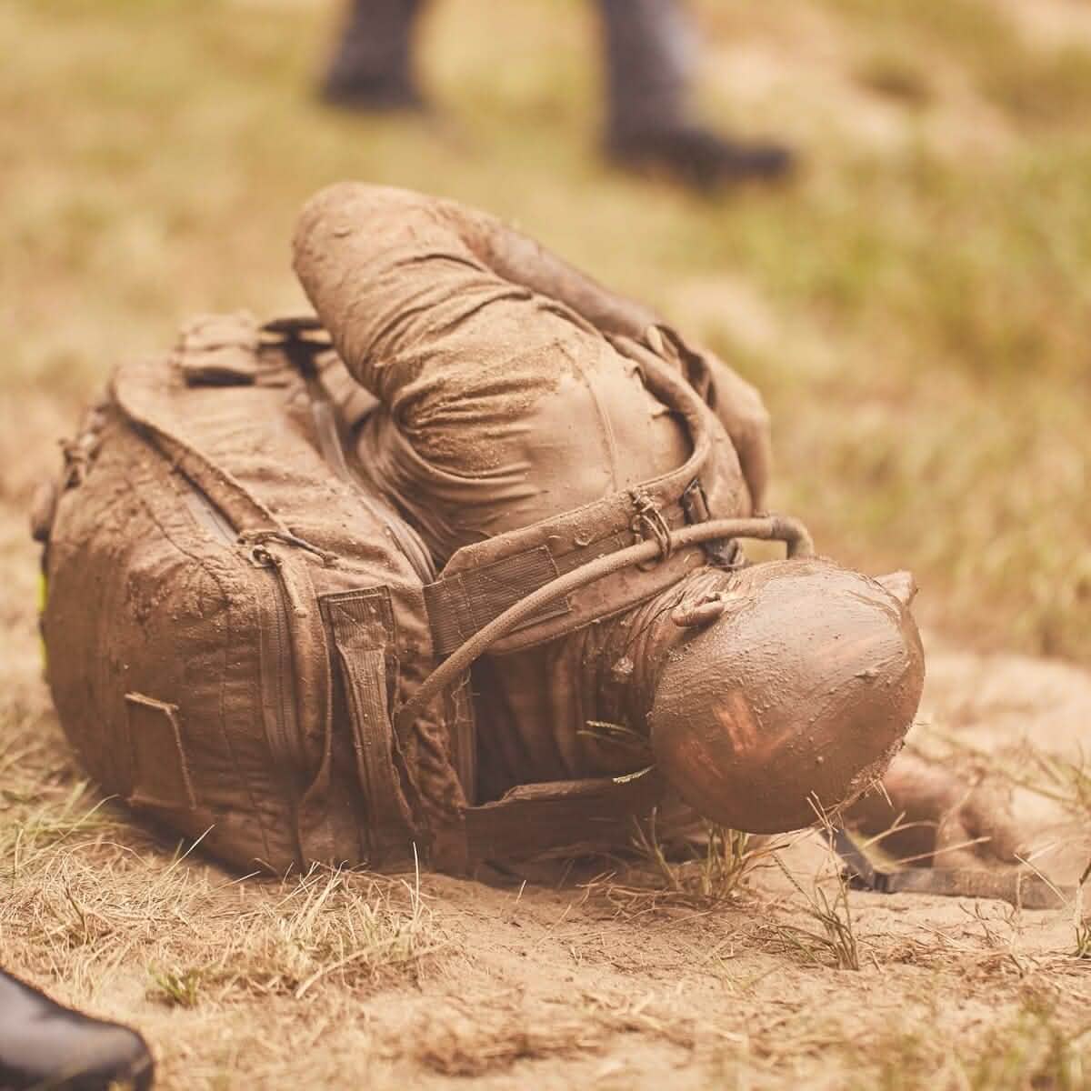 Amongst the grassy landscape, someone in a mud-covered uniform lies gripping their side, with the hose of a hydration bladder securely held by a Bladder Hose Retainer. Nearby, another person's legs can be seen, symbolizing the endurance associated with GORUCK events.
