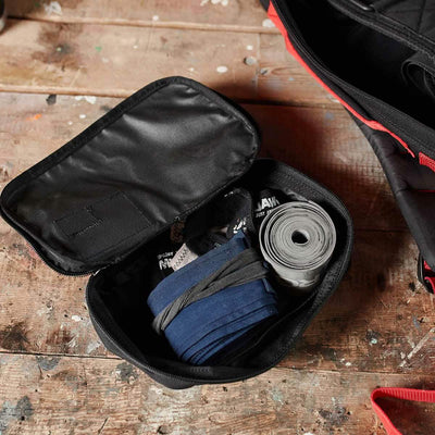 A black Shadow Pockets tool pouch from GORUCK lies on a wooden surface, showcasing its impressive organization. Inside, you'll find a coiled bicycle inner tube, blue tire levers, and a multi-tool neatly arranged within the shadow pockets. The pouch lid is open.