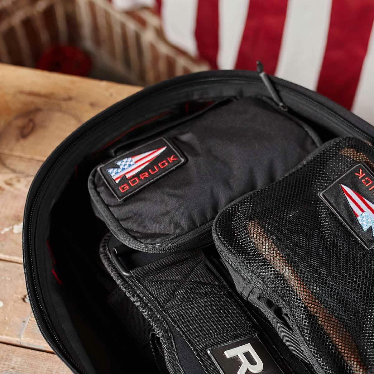 A close-up of a partially open backpack on a wooden surface shows the organization provided by the Shadow Pockets. Inside, one pocket features a patch displaying the American flag and the word GORUCK next to a folded flag. The use of durable Cordura ensures everything is held securely in place.