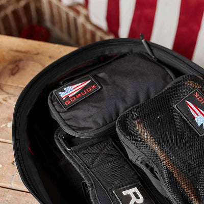 A close-up of a partially open backpack on a wooden surface shows the organization provided by the Shadow Pockets. Inside, one pocket features a patch displaying the American flag and the word GORUCK next to a folded flag. The use of durable Cordura ensures everything is held securely in place.