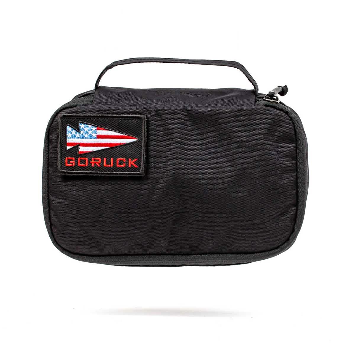 Introducing the GORUCK Shadow Pockets: a black travel pouch made from durable Cordura, complete with a carrying handle. It showcases a rectangular patch adorned with an arrow design and an American flag motif, highlighted by the word 'GORUCK' in red lettering—ideal for keeping your ruck organized on your adventures.
