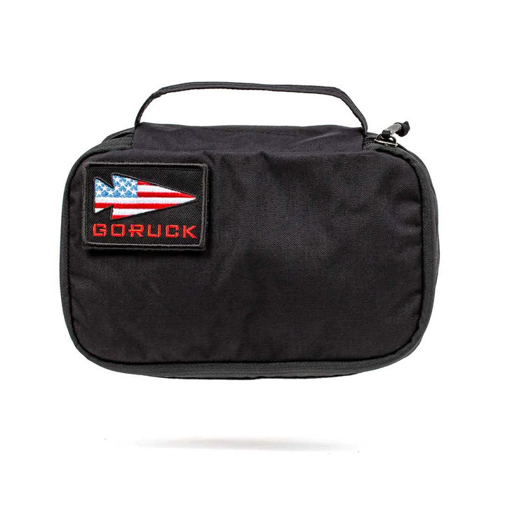 Introducing the GORUCK Shadow Pockets: a black travel pouch made from durable Cordura, complete with a carrying handle. It showcases a rectangular patch adorned with an arrow design and an American flag motif, highlighted by the word 'GORUCK' in red lettering—ideal for keeping your ruck organized on your adventures.