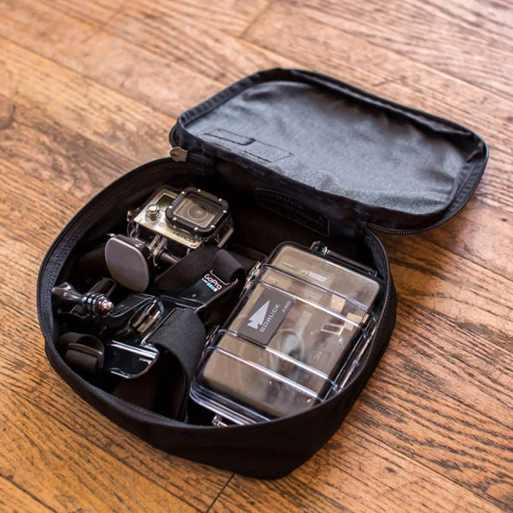 The wooden floor is home to an open black zippered case from GORUCK, featuring their innovative Shadow Pockets design. This durable Cordura case securely holds a small action camera, protective housing, and various accessories for seamless organization.