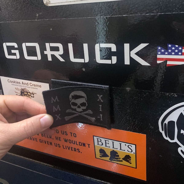 A hand is attaching a Patch with a skull and crossbones design onto a magnetic surface filled with various stickers, including one featuring the GORUCK logo next to an American flag. Other visible stickers are from Cookies And Creme and Bells Brewery.