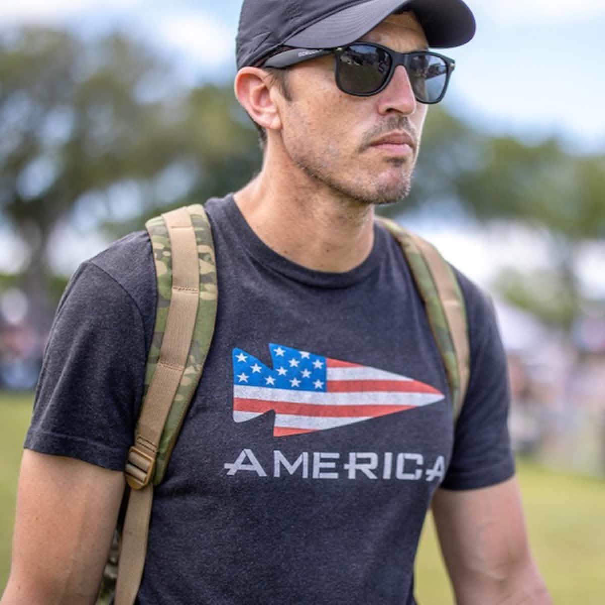 A man is wearing the America Tee - Tri-Blend by vendor-unknown, featuring an American flag design and "America" text. Paired with sunglasses and a black cap, he carries a camouflage backpack while standing outdoors with blurred trees behind him. This stylish shirt is proudly printed in the USA.
