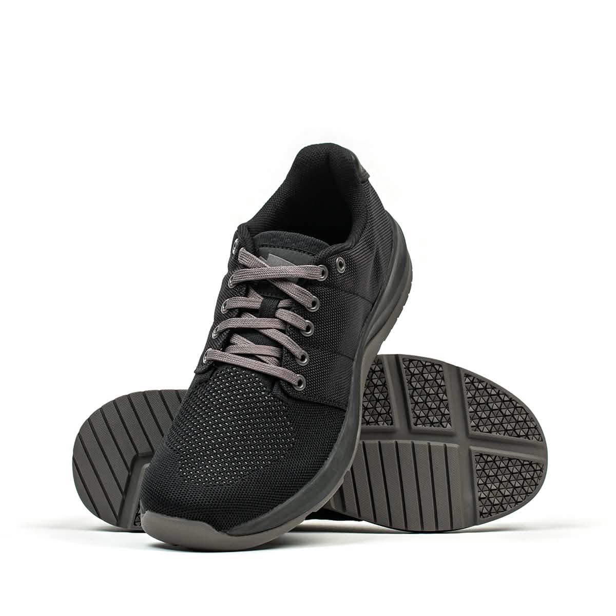 GORUCK's Men's Ballistic Trainers in Black + Black + Charcoal are on display. One sneaker is positioned upright, prominently showcasing the mesh and CORDURA® Ballistic Nylon features, while the other shoe is flipped sole-up, revealing its textured rubber finish designed for functional fitness activities.