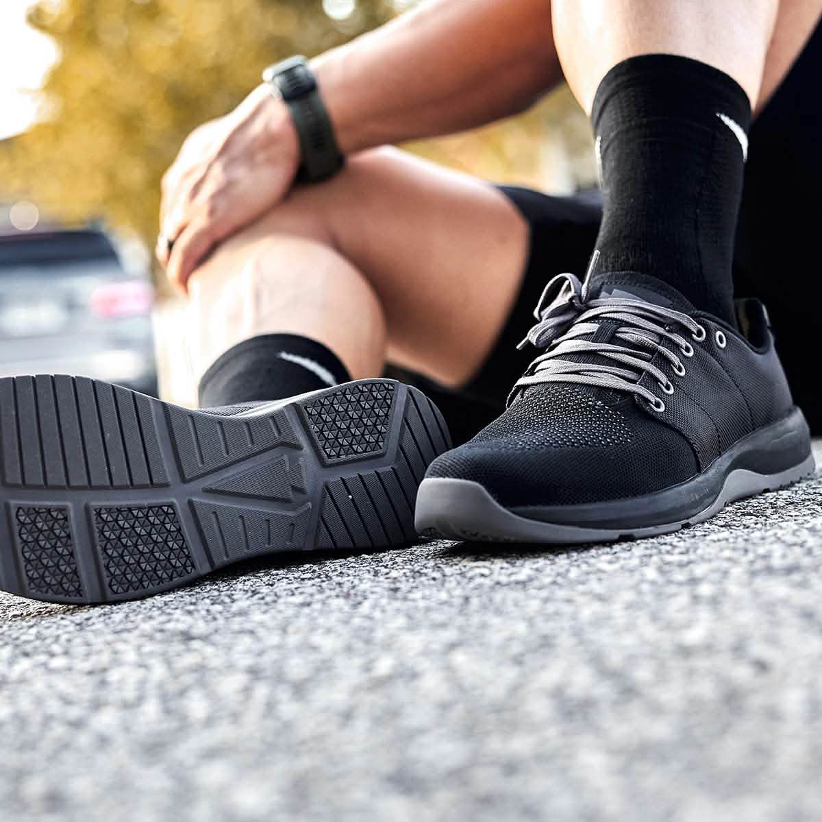 A person sits on the pavement wearing GORUCK's Men's Ballistic Trainers in Black, Black, and Charcoal, complemented by black socks. The focus is on these functional fitness shoes, highlighting their design and soles made from durable CORDURA® Ballistic Nylon. In the background, blurred greenery and a parked car are visible.