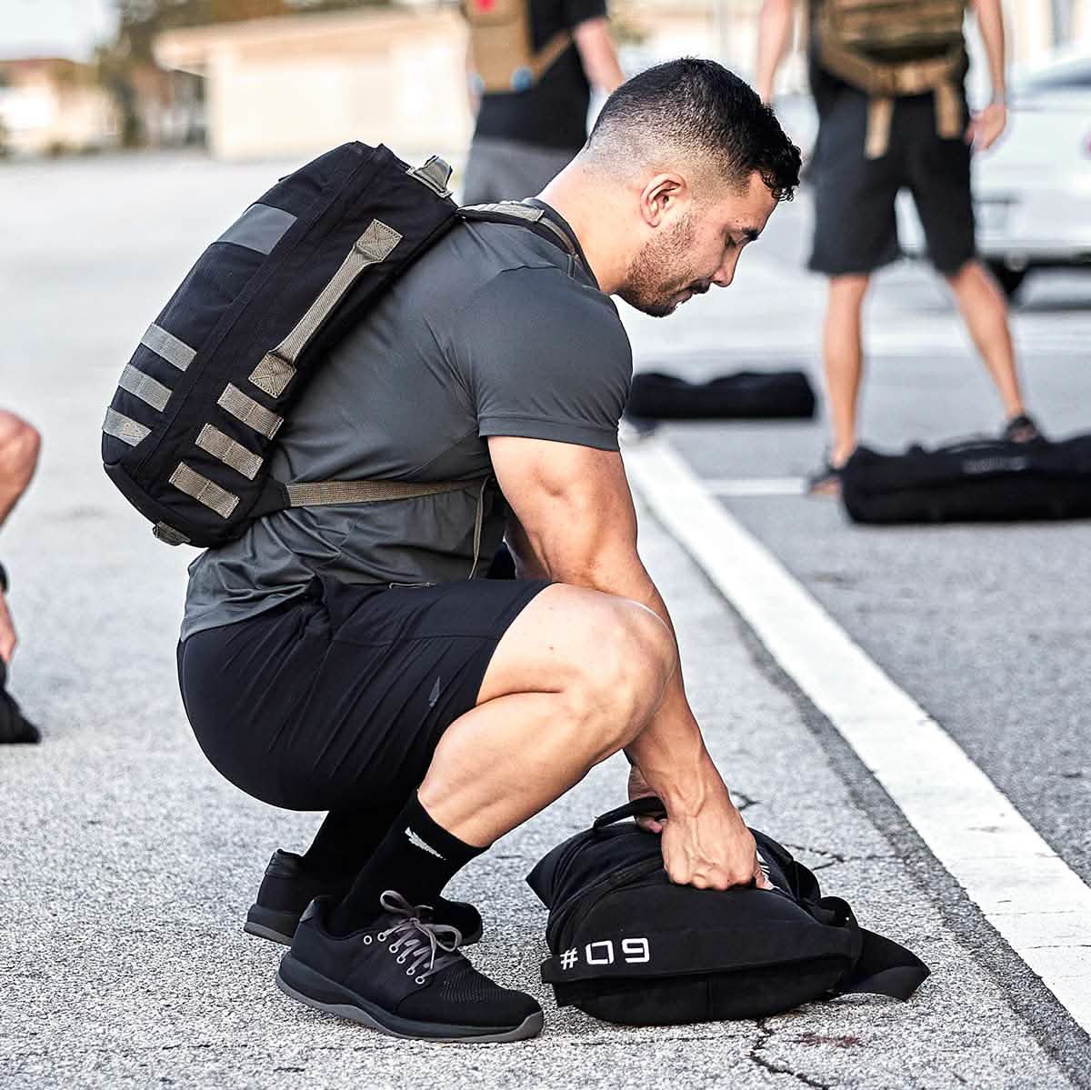 A man in athletic wear and a backpack is crouching on a pavement, sporting GORUCK Men's Ballistic Trainers in Black + Black + Charcoal. He's handling a black bag marked #09, while others with backpacks can be seen in the background, indicating an outdoor training environment focused on endurance and strength.