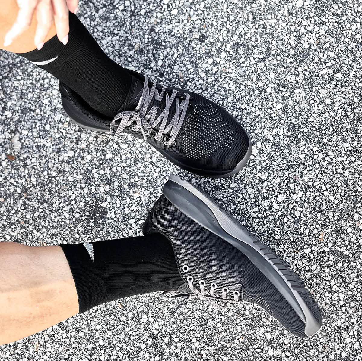 A person is positioned on a speckled gray surface, donning the Men's Ballistic Trainers - Black + Black + Charcoal by GORUCK. The trainers, crafted from durable CORDURA® Ballistic Nylon with gray laces, are specifically designed for 3X Support functional fitness. One foot is slightly lifted, highlighting how well these shoes pair with their black socks. Only the lower legs are visible.