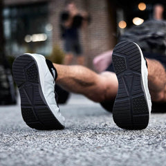 Close-up of the soles of Ballistic Trainers - Slick - Lunar Rock + Charcoal as someone lies on their back on a rough outdoor surface.