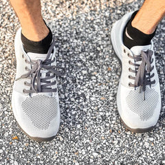 Person wearing Ballistic Trainers - Slick - Lunar Rock + Charcoal and black socks stands on textured outdoor pavement, showcasing workout shoes with 3X Support for superior stability and comfort.