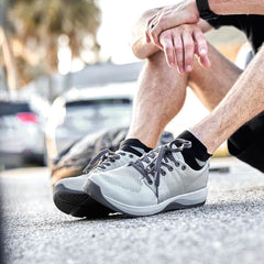 A person sits on the pavement wearing Ballistic Trainers - Slick - Lunar Rock + Charcoal, hands on knees, with cars visible behind.