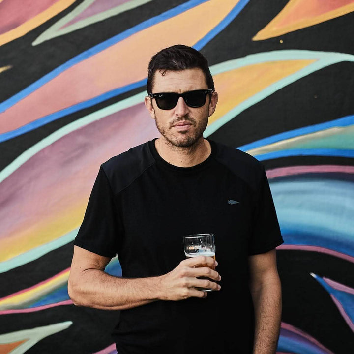 A man wearing sunglasses holds a glass of beer in front of a colorful, abstract mural. Dressed in the GORUCK Men’s Commando Tee - Merino Wool, the vibrant background enhances his style with its swirls and brushstrokes in various colors.