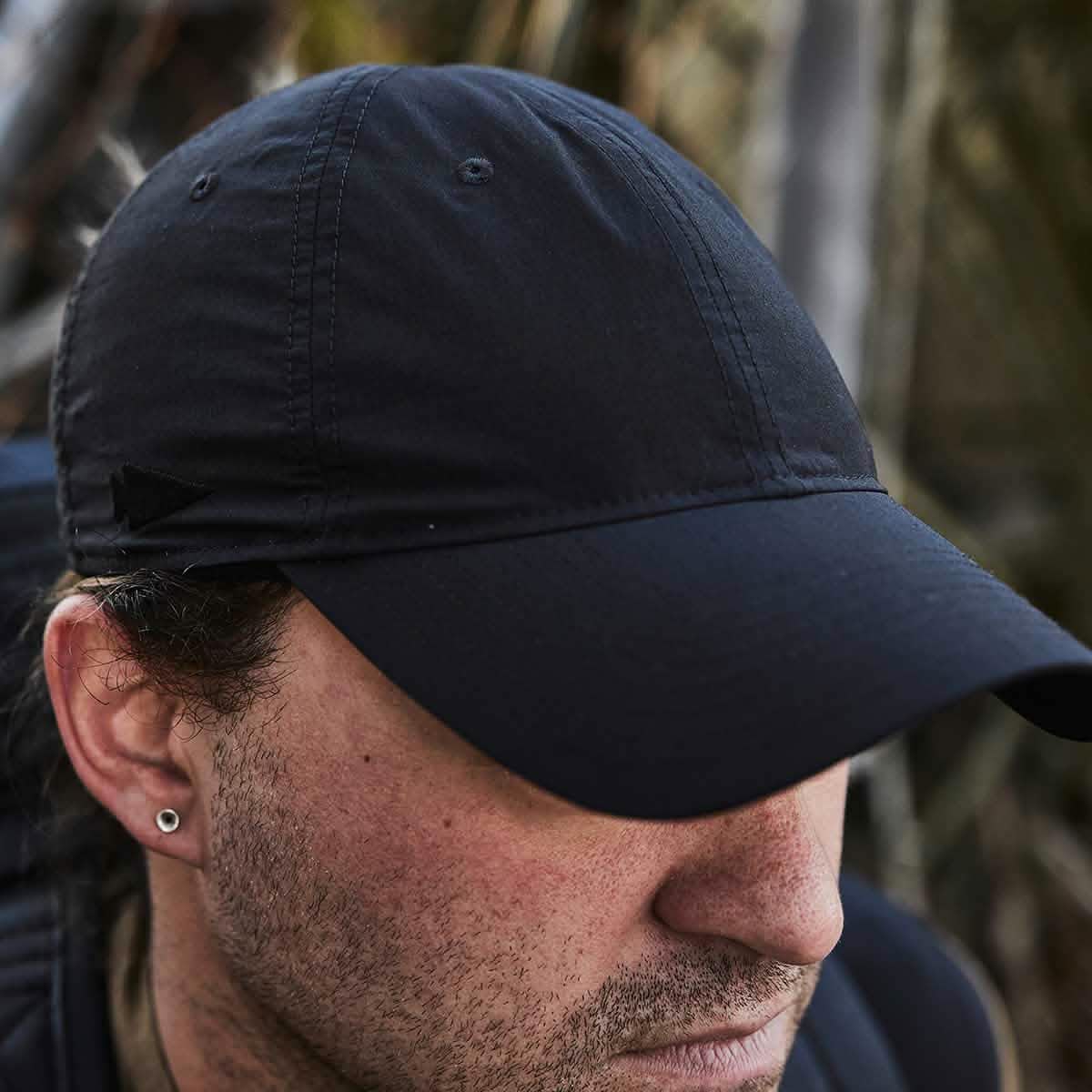 A person wearing the GORUCK Performance TAC Hat - Slick in TOUGHDRY fabric glances downward, partially hiding their face. An earring dangles from their left ear, while the background of indistinct foliage emphasizes the hat’s sweat-wicking properties for active individuals.