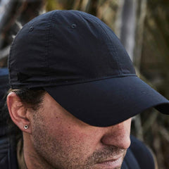 A person wearing the GORUCK Performance TAC Hat - Slick in TOUGHDRY fabric glances downward, partially hiding their face. An earring dangles from their left ear, while the background of indistinct foliage emphasizes the hat’s sweat-wicking properties for active individuals.