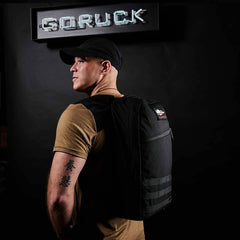 A man wearing a black cap and brown shirt models the GORUCK Bullet Ruck Laptop - 16L, highlighting its sleek design with a laptop compartment. He stands against a dark background illuminated by a GORUCK sign, looking to the side and revealing an arm tattoo.