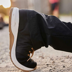 A close-up of a person wearing black pants and GORUCK's Men's Ballistic Trainers in Black, White, and Coyote, lying on a sandy surface. The setting sun casts a warm glow in the background.