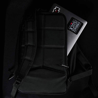 A black GORUCK Bullet Ruck Laptop - 16L rests on a dark surface, with a laptop partially visible inside. The laptop features a sticker that says "EMBRACE THE SUCK" and displays a red star. The backpack includes padded sections, lumbar support, and adjustable straps for added comfort.