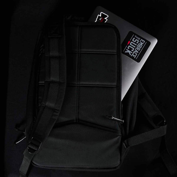 A black GORUCK Bullet Ruck Laptop - 16L rests on a dark surface, with a laptop partially visible inside. The laptop features a sticker that says "EMBRACE THE SUCK" and displays a red star. The backpack includes padded sections, lumbar support, and adjustable straps for added comfort.