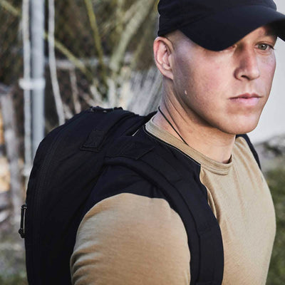 A person wearing a black cap and a tan shirt carries the GORUCK Bullet Ruck Laptop - 16L, which offers lumbar support. They are outdoors, with a blurred background featuring a fence and greenery.