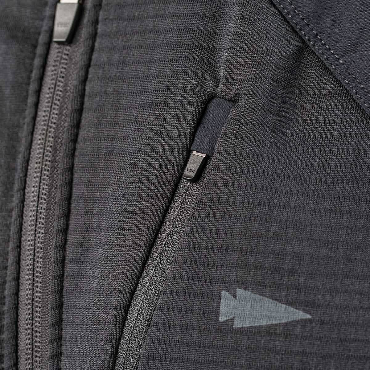 Close-up of the GORUCK Men's Full Zip - Polartec Grid Fleece jacket in a gray-textured fabric, showcasing a partially closed zipper. Above it, there’s a small zippered pocket with a pull tab. On the right side of the ToughDry Fabric, a subtle arrow-shaped design adds both durability and style to this fleece-lined jacket.