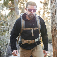 As they hike through the forest, wearing sunglasses and a backpack, they bask in the sunlight filtering through the trees. Dressed in GORUCK's Men’s Full Zip - Polartec Grid Fleece jacket and tan pants, they embrace the warm, adventurous atmosphere.