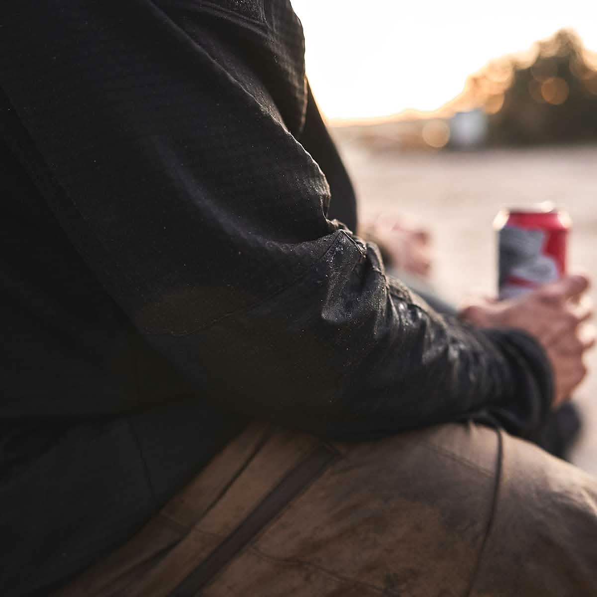 A person wearing a Men’s Full Zip - Polartec Grid Fleece by GORUCK in black and paired with brown pants sits outdoors, holding a red and white can. The background is blurred, suggesting a natural setting at sunset.