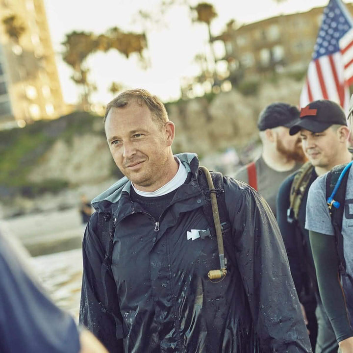 A group of people walking outdoors under the trees, with one person in the foreground wearing a raincoat and carrying a small axe. Amidst them, someone adjusts their GORUCK Bladder Hose Retainer. In the background stands a building and another person proudly holding an American flag.