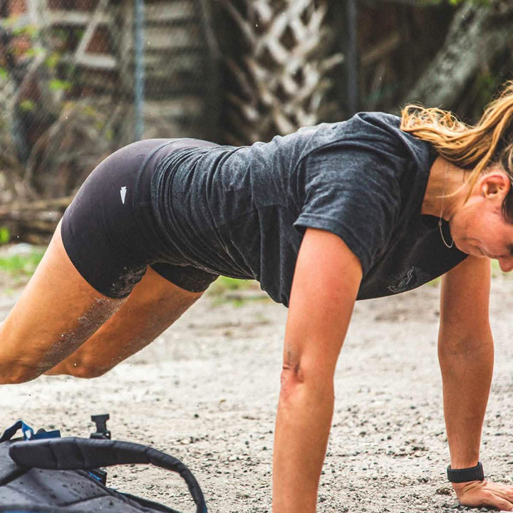 A person exercises outdoors, holding a plank position on sandy ground. Clad in GORUCK Women’s Squat Shorts - ToughFlex and a shirt, sand flecks dot their legs. Nearby rests a water bottle, with a fence and lush greenery providing the backdrop.