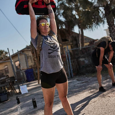 A person wearing sunglasses and a Seek Pain t-shirt lifts a heavy bag above their head during an outdoor workout in Women’s Squat Shorts made of ToughFlex by GORUCK. Another person is bent over in the background, with palm trees and a fence visible under the clear sky.