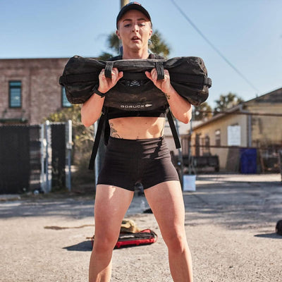 A person wearing a cap and athletic attire, including GORUCK Women’s Squat Shorts made from ToughFlex Fabric, lifts a heavy sandbag outdoors against the backdrop of buildings and a fence on a sunny day.