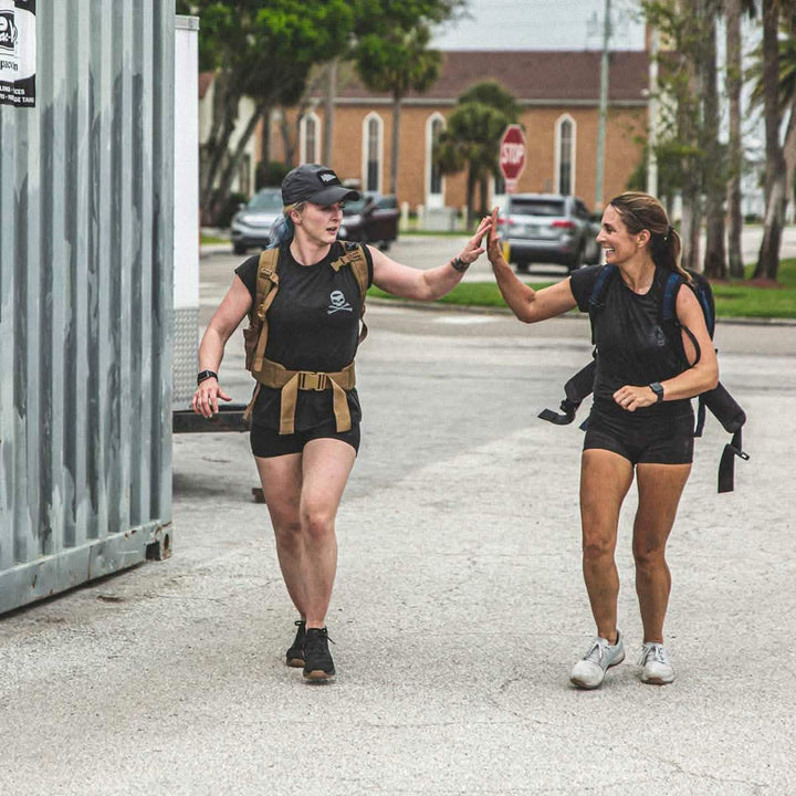 Two women in athletic gear high-five while walking outdoors. One is clad in a cap and tactical vest, while the other showcases a backpack paired with GORUCK's Women’s Squat Shorts made from ToughFlex Fabric. They're surrounded by shipping containers, cars, trees, and a building in the background.