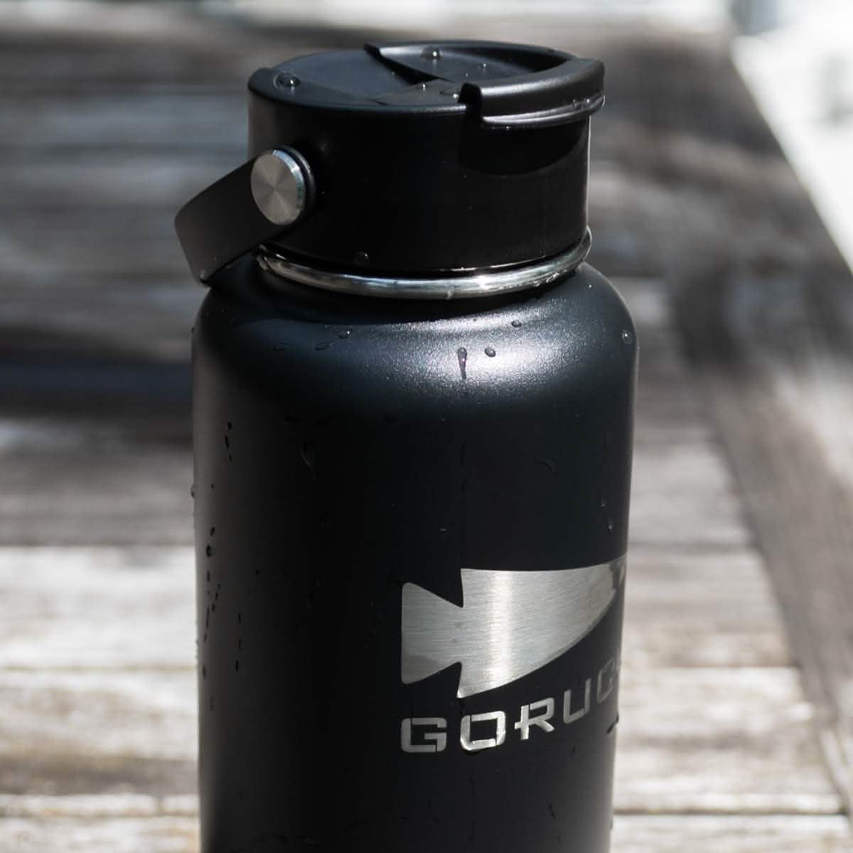 On a wooden table sits a Water Bottle - GORUCK x RTIC, designed in black stainless steel with a silver logo. The bottle displays light water droplets on its surface and features a screw-top lid with a handle, emphasizing its double-wall insulated design.