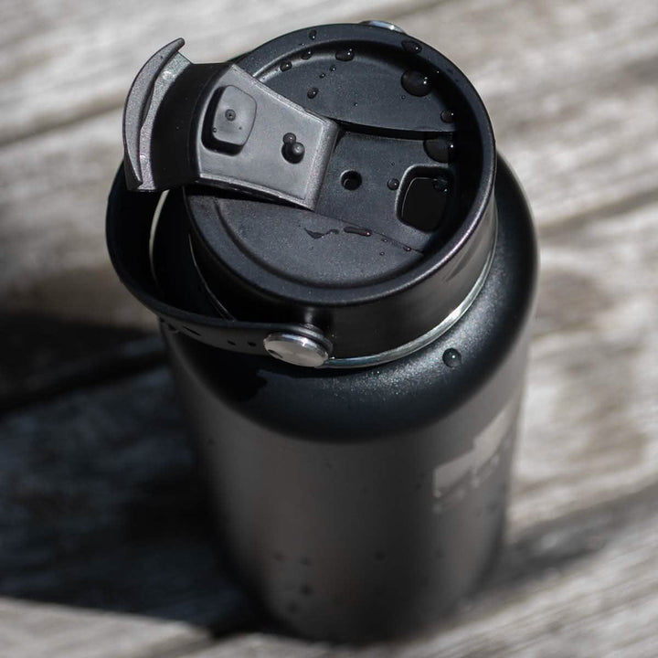 A black Water Bottle - GORUCK x RTIC, featuring a double wall insulated stainless steel body and a sleek design similar to RTIC bottles, rests on a weathered wooden surface. Water droplets cling to its closed flip-top lid and body, suggesting it was recently used or filled with cold liquid.