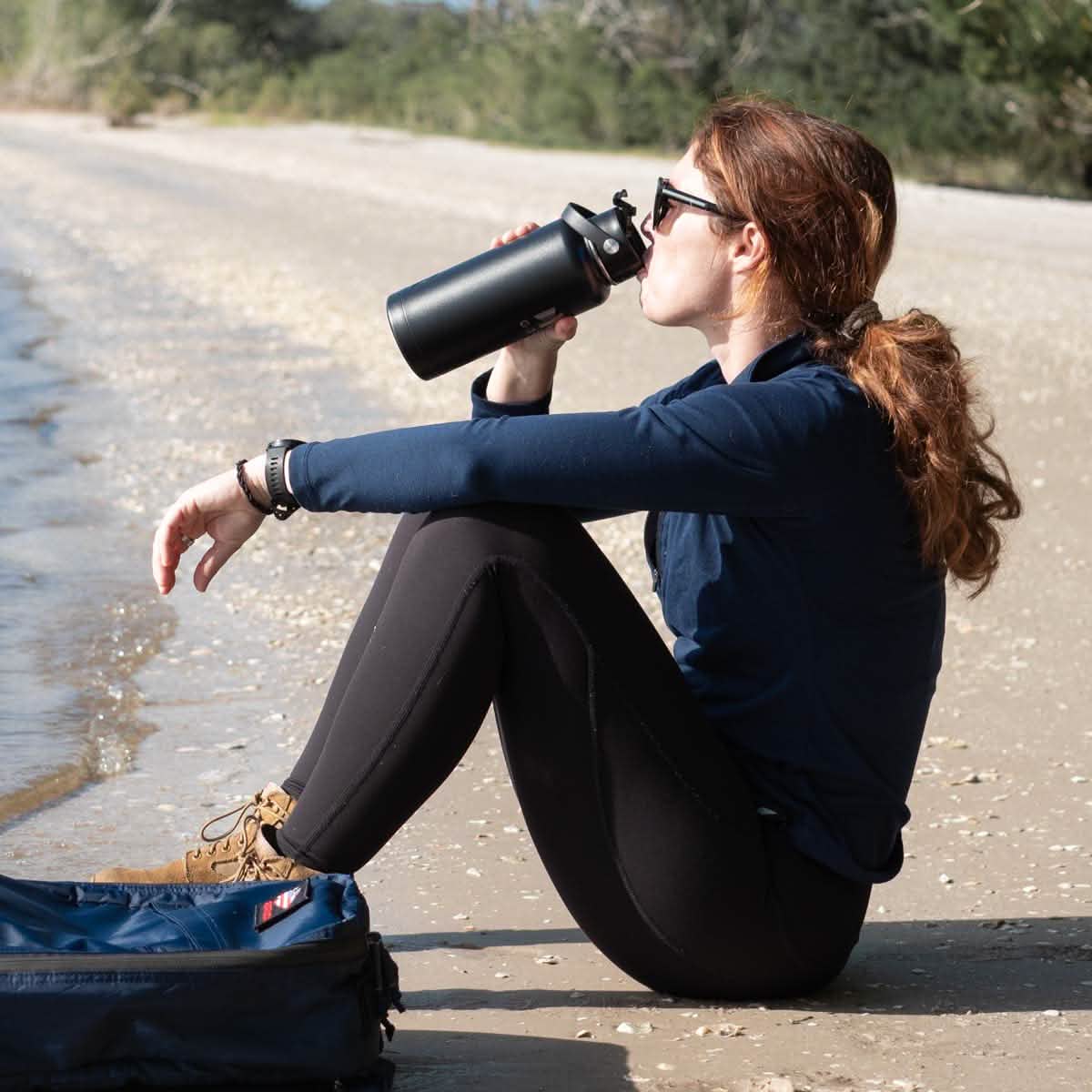 A person with long hair wearing sunglasses, a navy jacket, and black leggings sits on a sandy beach near the water, sipping from a sleek stainless steel Water Bottle - GORUCK x RTIC by GORUCK. A blue backpack is beside them. The scene is calm and sunny.