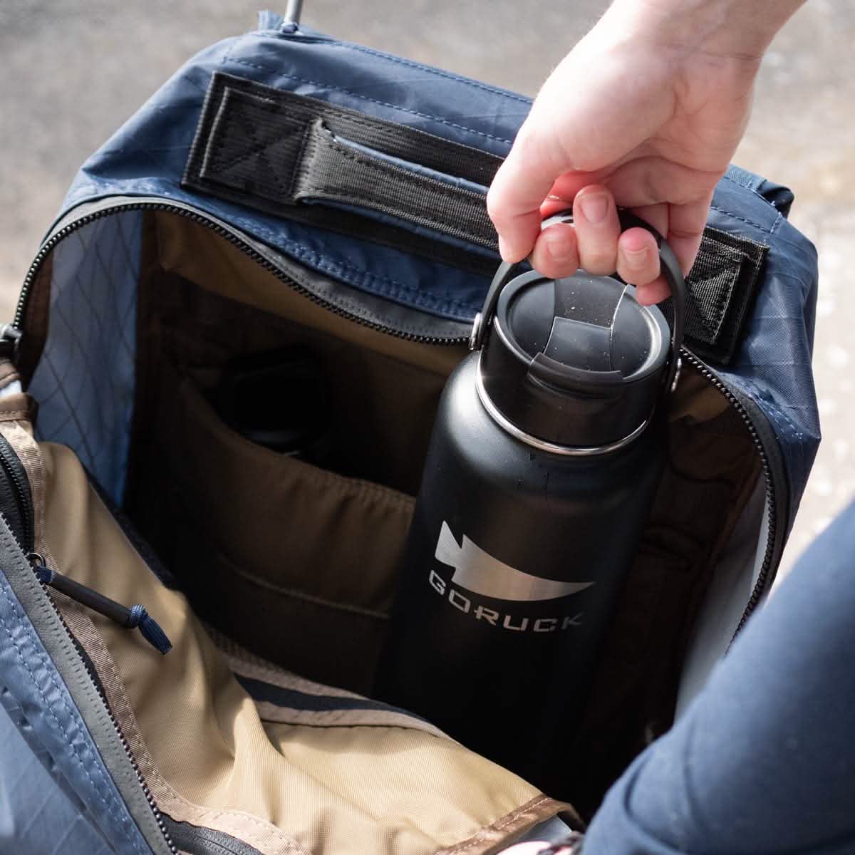 A person places a black, double-wall insulated GORUCK x RTIC water bottle into a blue backpack, revealing its beige interior with multiple compartments.