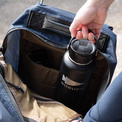 A person places a black, double-wall insulated GORUCK x RTIC water bottle into a blue backpack, revealing its beige interior with multiple compartments.