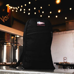 A black Bullet Ruck Laptop - 16L from GORUCK is placed on a surface in a dimly lit room adorned with string lights overhead. Industrial elements are visible in the background, emphasizing its sleek design. The backpack features lumbar support and a compartment for a laptop, enhancing its functionality.