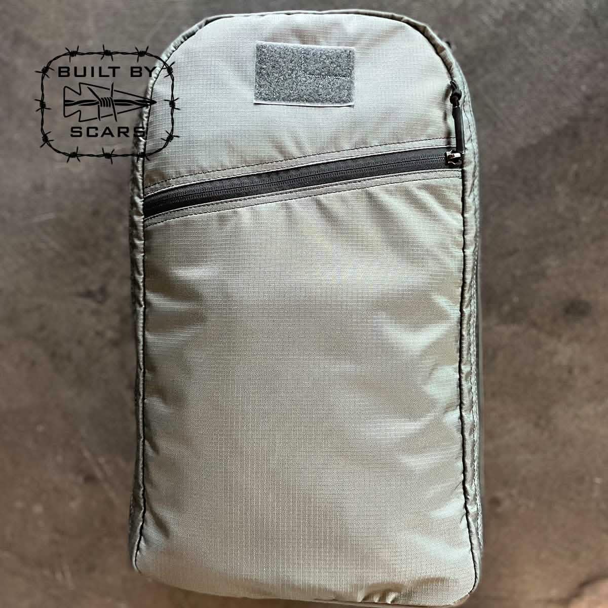 The Bullet 15L Robic Nylon backpack in gray, manufactured by GORUCK, is showcased against a textured background. It includes a front zippered pocket and prominently displays the 