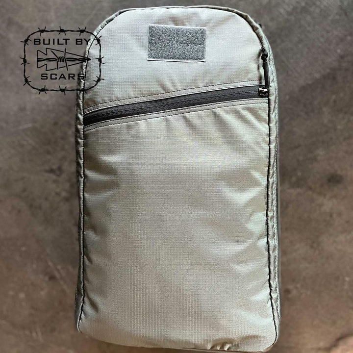 The Bullet 15L Robic Nylon backpack in gray, manufactured by GORUCK, is showcased against a textured background. It includes a front zippered pocket and prominently displays the "Built by SCARS" logo featuring barbed wire and a shark on the left side.