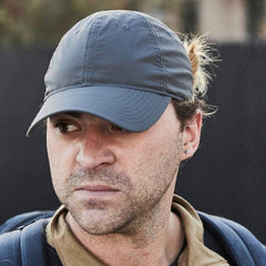 A person wearing a GORUCK Performance TAC Hat - Slick - TOUGHDRY and a brown jacket looks to the side. The individual, sporting a backpack and an earring, has their hair tied back. The background is blurred, emphasizing the person's contemplative expression.